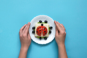 Funny sandwich for children in form of ladybug made of fresh tomato, salad, cheese, sausage, bacon and bread on blue background. Instruction how to make creative food art breakfast for kids.