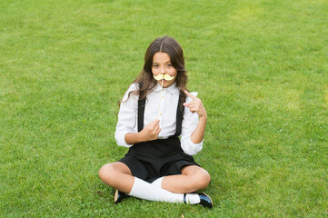 Funny party concept. Cute mustache. Small girl hold fake mustache on face. Happy childhood. Little child school uniform. Back to school. Small schoolgirl sit on green grass. Fashion accessory