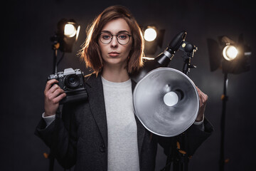 Fototapeta premium Portrait of a beautiful redhead female photographer holds a digital camera and looking on the camera. Studio shot on dark background with lighting equipment