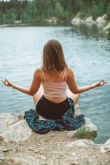 Young woman practicing yoga in the nature, sitting on the stone back to the camera and meditation, lake, active life concept, summer time