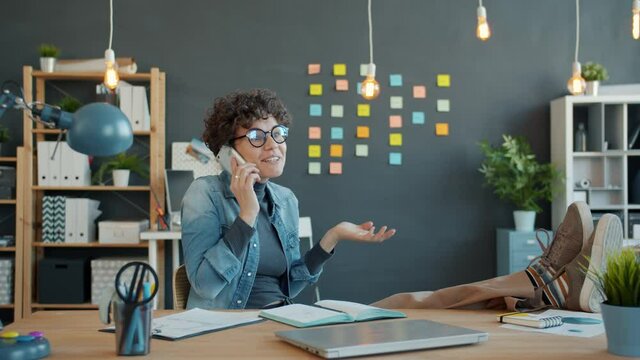 Joyful young woman is chatting on mobile phone talking and gesturing at desk in office having fun with enjoyable conversation. People and devices concept.