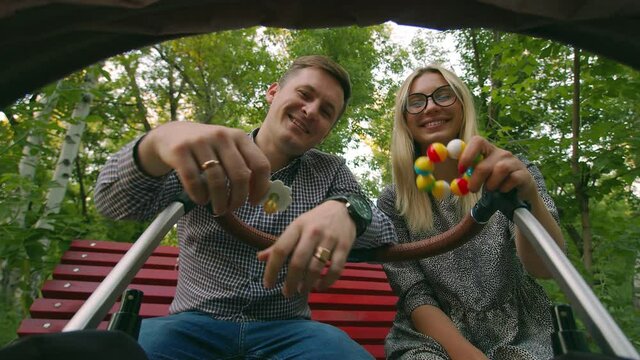 A happy young family looks at the baby in the stroller, the mother shakes the toy, the father brings the pacifier. Laughing dad puts a pacifier in the camera, mom shakes the toy