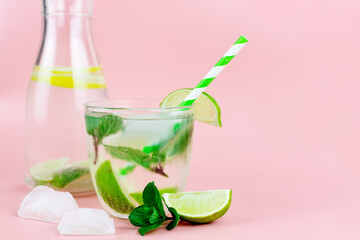A carafe of refreshing water with lemon or lime and ice and in the foreground a glass with a cocktail on a pink background. Summer cold drinks