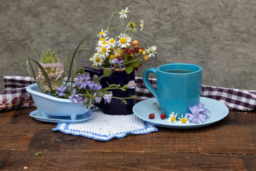 On the table is a bouquet of daisies and wild strawberries, a cup of tea, on a saucer lies wild strawberries daisy flowers and chicory. Nearby are chicory flowers.
