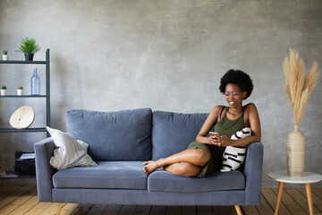 Happy african American young woman sit relax on cozy couch happy to move to new apartment. Smiling black girl rest on comfortable sofa in living room dreaming