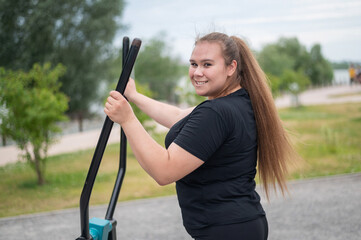 Beautiful smiling overweight young woman does fitness on an ellipsoid simulator outdoors. Fat girl...
