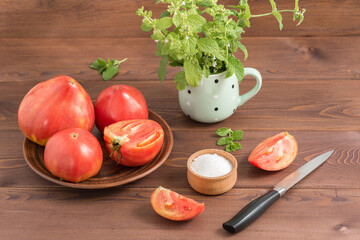 Large ripe juicy country pink tomatoes, a salt shaker with salt and a knife on a wooden table. In the mug - bunch of Melissa