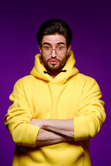 A young man of 25-30 years in glasses and a yellow sweatshirt emotionally poses on a purple background. 