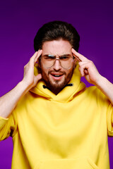 A young man of 25-30 years in glasses and a yellow sweatshirt emotionally poses holding his head on a purple background.