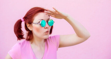 Audacious young woman in pink shirt and blue round sunglasses looking for something, holding palm near forehead like visor protecting eyes from sunlight. Pink background. Copy space