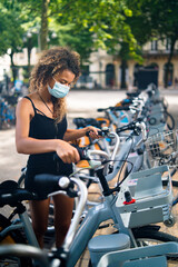 Fototapeta premium Young woman wearing mask against coronavirus using city rental bicycles