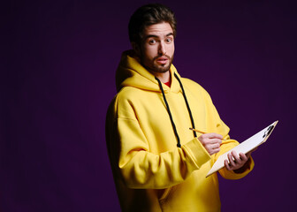 A young man of 25-30 years in glasses and a yellow sweatshirt holding a clipboard with clip in one hand and a pencil in the other