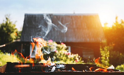 Burning firewood in a homemade barbecue on background of green foliage and village house at sunset. Firewood, fire and smoke with warm back light