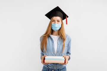 graduate girl in a medical mask on her face and a graduation hat on her head, with books in her hands, stands on a white background. Graduation, distance education, coronavirus