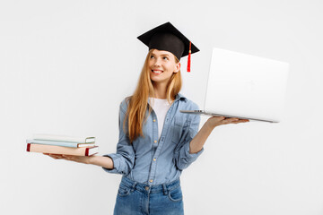 thoughtful, focused graduate in a graduation cap on her head, with a laptop and books in her hands,...