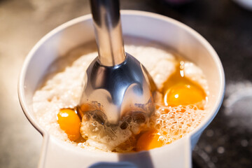 Whipping the eggs and flour in a bowl with a hand blender to prepare a recipe. Prepare a homemade cake at home.