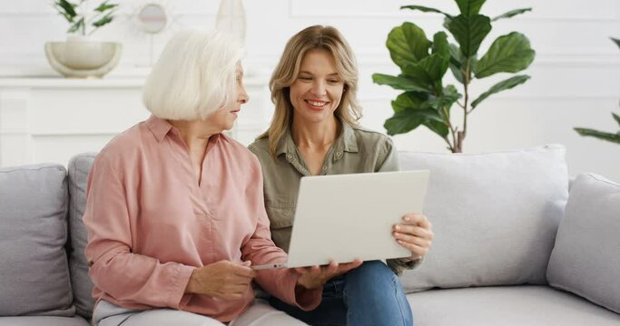 Beautiful Caucasian adult daughter showing something on laptop to old gray-haired mother and discussing that. Women of different ages sitting on couch, using computer at home. Senior woman talking.