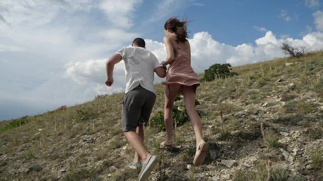 A young couple holding hands runs up a rocky slope to meet lush clouds. A guy in a white T-shirt and a girl in a pink dress with her hair flying in the wind run down the hill