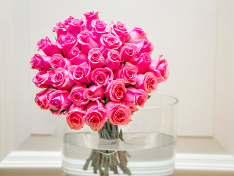 Large Bouquet Of Two Dozen Pink Roses In Large Glass Vase With Water Against White Painted Cove