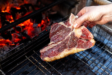 Close-up man hand putting big tasty juice raw t-bone beaf steak on grill at barbecue with bright hot burning charcoal at brazier. BBQ meal cooking background