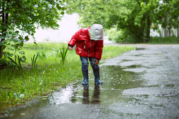 a three-year-old boy in a red jacket with a hood stands in a puddle on the pavement and touches his wet pants with his hand.
lmage with selective focus