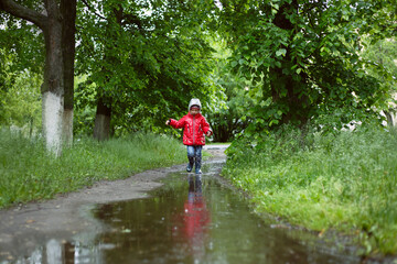 a boy in a red jacket with a hood and blue boots runs in the rain among the trees in a deep puddle, water splashes fly.
lmage with selective focus
