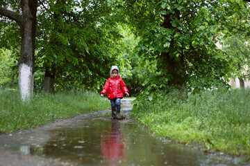 little boy in a red jacket with a hood and blue boots walks in the rain among the trees in a dirty puddle, looks down.
lmage with selective focus
