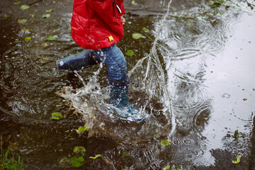 a boy in blue jeans, patterned boots and a red jacket during the rain jumps in a large puddle, splashes fly.
lmage with selective focus