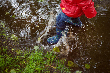 a boy in blue jeans, patterned boots and a red jacket steps into a large puddle during the rain.
lmage with selective focus