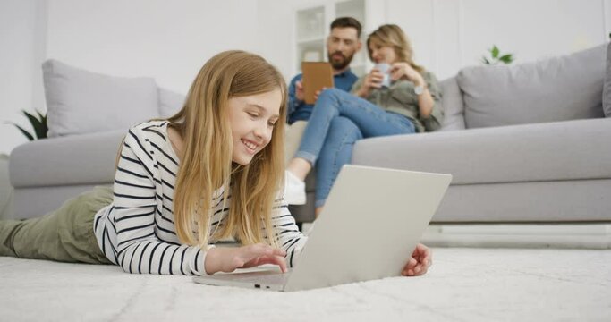 Caucasian happy kid lying on floor and typing on keyboard of laptop. Small cute girl playing on computer at home. Parents sitting on couch and talking on background. Mother and father rest.