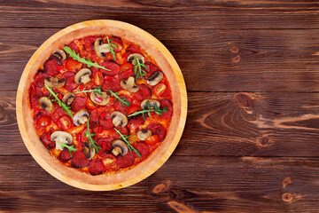 Whole pizza hot with spicy salami, arugula, cherry tomatoes, mushrooms and texas spice mix, on a round wood plate which is on wooden rustic table, top view and copy space