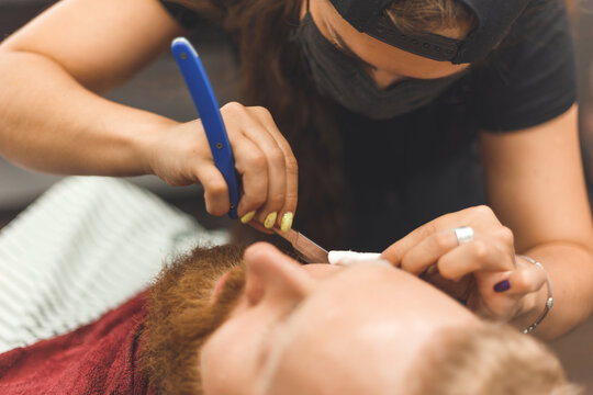 Barber woman shaves beard with a straight razor. Hairdresser equipment. Selective focus. Master in face mask