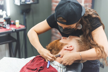 Barber woman shaves beard with a straight razor. Hairdresser equipment. Selective focus. Master in...