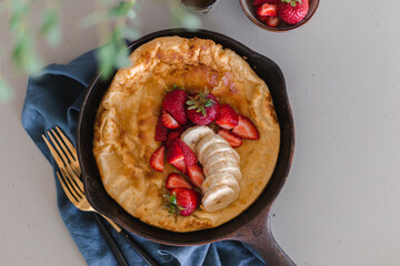Dutch baby pancake baked until golden brown in cast iron skillet and topped with strawberries, banana and maple syrup. Weekend brunch meal. Sunday breakfast. Simple recipe. Fluffy battered pancake.