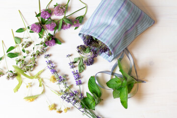 Cotton sachet bag with dry red clover flowers and fresh herbs of mint, linden, lavender, oreganum on white wooden background