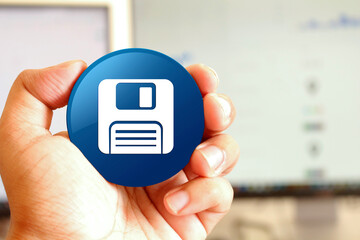 Floppy disk icon blue round button holding by hand infront of workspace background