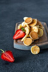Trendy home breakfast with tiny pancakes (mini pancakes) with strawberries and mint on a wooden board on a dark background. Food Background. Mini cereal pancakes.