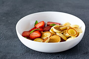 Trendy home breakfast with tiny pancakes (mini pancakes) with strawberries and mint in a bowl on a dark background. Food Background. Mini cereal pancakes.