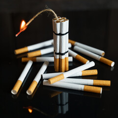 Cigarettes, in the form of sticks of dynamite with a burning wick, stand among the scattered cigarettes on a black reflective surface.