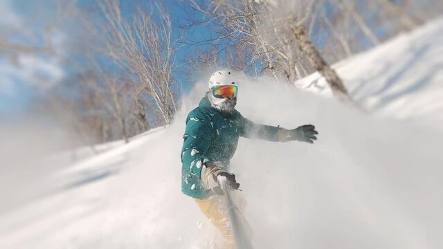 Concept of extreme, sport, winter, freeride, snowboarding. Man riding on snowboard with selfie stick in his hand between trees on slow motion. Guy doing snow splash rising on magnificent sunny day.