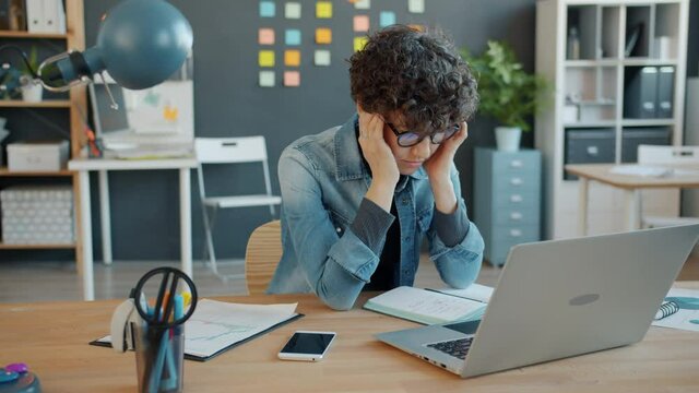 Tired businesswoman is massaging head feeling exhausted working with computer in office at desk. Hard-working youth, technology and health concept.