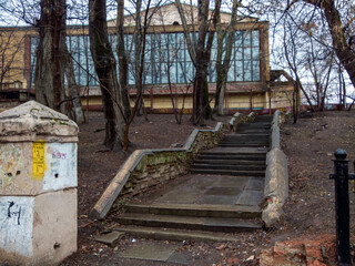 Fototapeta na wymiar An old staircase with wet steps leading to an abandoned Soviet building, through bare trees, behind a fence. Palace of Culture of the Hammer and Sickle Factory