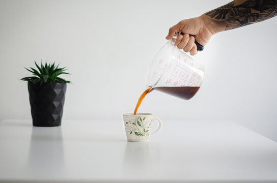 Mano de hombre vertiendo caf&eacute; reci&eacute;n hecho en una taza sobre una mesa blanca, en segundo plano una maseta negra con una planta. Proceso de preparaci&oacute;n de caf&eacute; colombiano. 