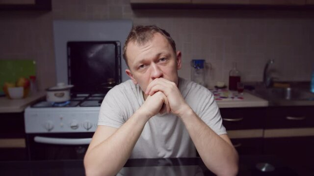 A depressed man is sitting at the kitchen table in the evening, looking directly at the camera, camera movement