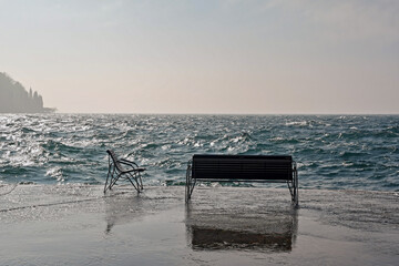 Backlight view of the lakeside with high waves, empty benches and a cape on the horizon, Lake Garda, Italy