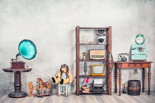 Antiques still life with old gramophone, Teddy bear, rocking horse, collection of media and writers devices, outdated cash register, clock front concrete wall background. Vintage style filtered photo