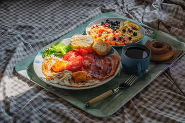 Breakfast at bed in a serving tray. Very high contrast scene with a plate of fried eggs and bacon, another plate with slices of fruit a donut and a cup of coffee.