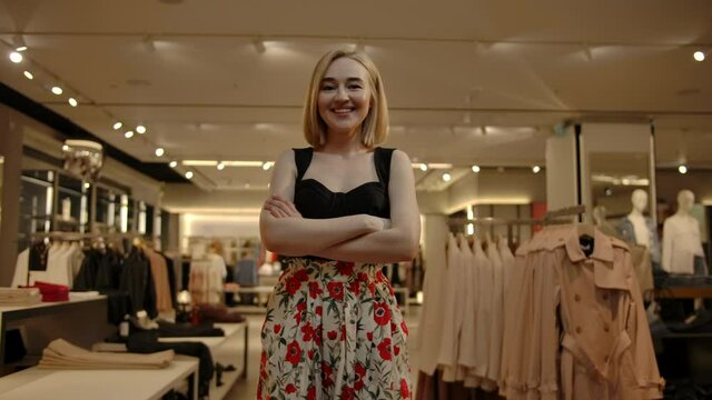 Confident and pretty beautiful woman standing with crossed arms indoor of fashionable clothes store. Smooth dolly camera movement. Portrait of smiling blondie during shopping.