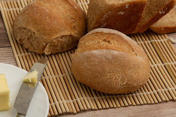 close up image of home made whole wheat bread loaves fresh out of oven. They are cooling on a bamboo mat on wooden table. A butter knife with butter on it is there to put delicious spread on a slice.