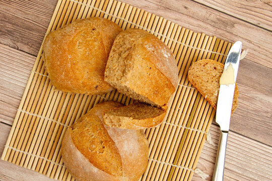 close up image of home made whole wheat bread loaves fresh out of oven. They are cooling on a bamboo mat on wooden table. A butter knife with butter on it is there to put delicious spread on a slice.